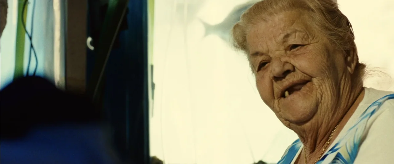 Reality (2023) movie still: closeup — An interior space faces a large glass water tank with a swimming fish visible. An elderly woman sits in front…