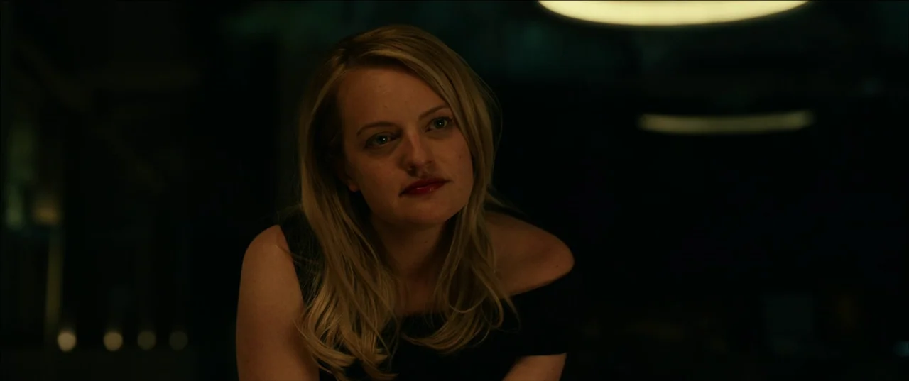 The Invisible Man (2020) (2020) movie still: closeup — A woman sits in a dark, out-of-focus modern lounge environment with soft illumination. She…