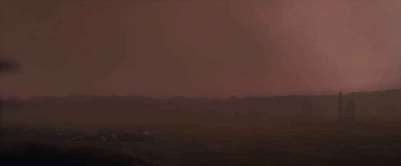 Ad Astra (2019) movie still: establishing — A desolate, hazy desert landscape under a thick, reddish-brown sky features distant jagged mountains.…