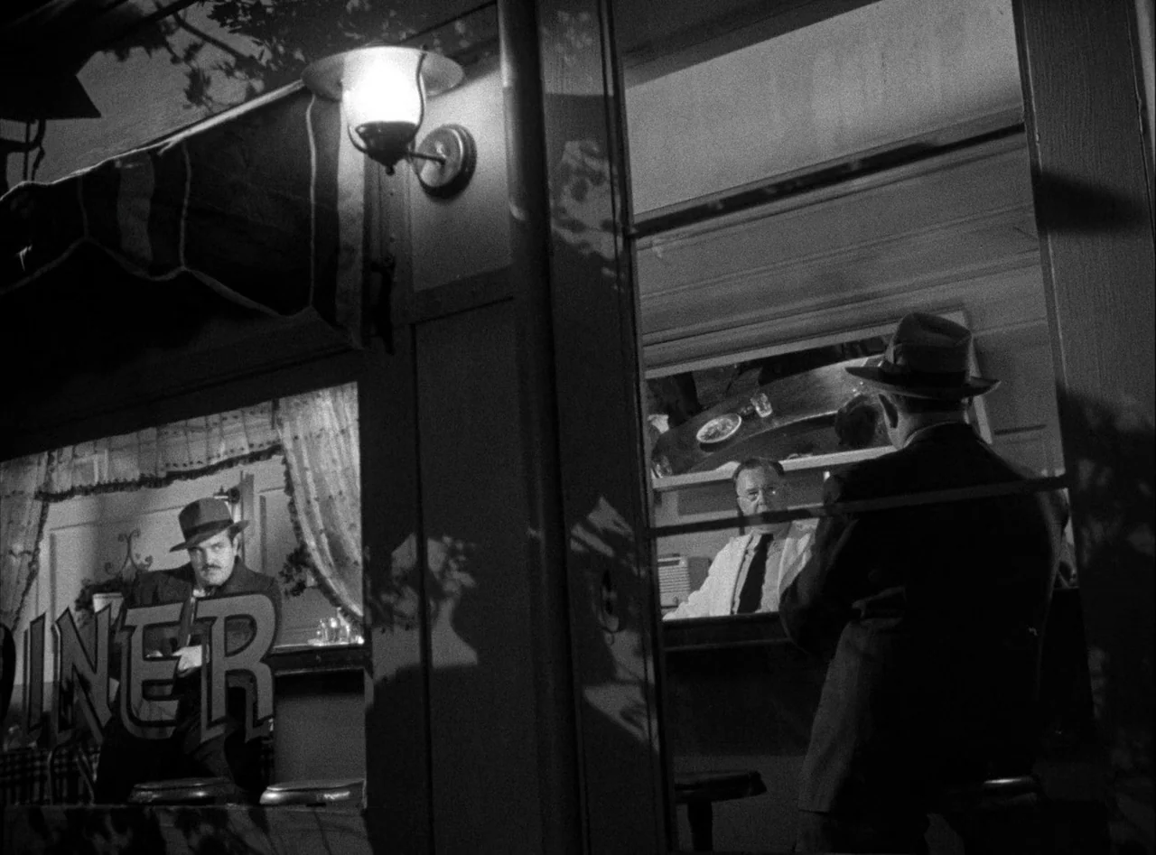 The Killers (1946) movie still: medium — A dark diner exterior at night features a large glass window pane reflecting street light. Two men sit…
