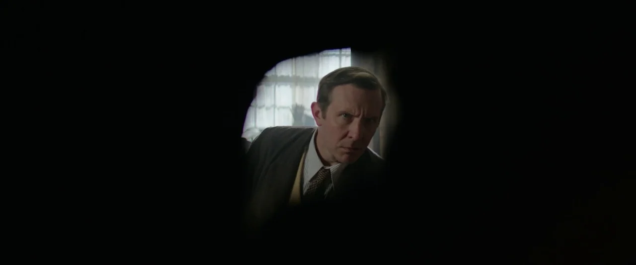 Nandor Fodor and the Talking Mongoose (2023) movie still: medium — A man is viewed through a dark, irregular hole in a wall or door. The man peers…