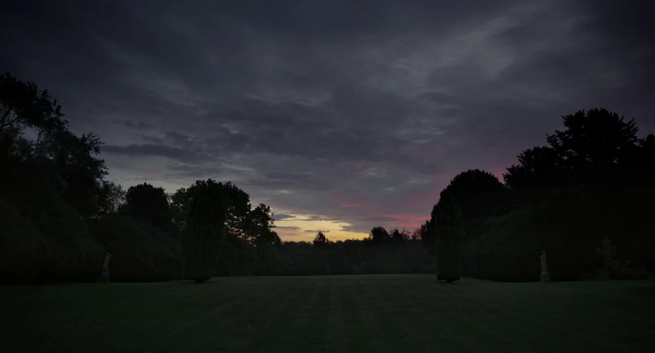 Flux Gourmet (2022) movie still: establishing — A vast, manicured lawn stretches toward a tree line under a dark cloudy sky. Faint pink and amber…