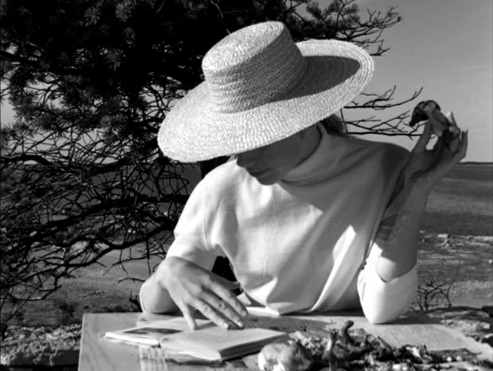 Persona (1966) movie still: medium — A woman wears a large straw hat while sitting at a table outdoors. She reads a book with her head down while…