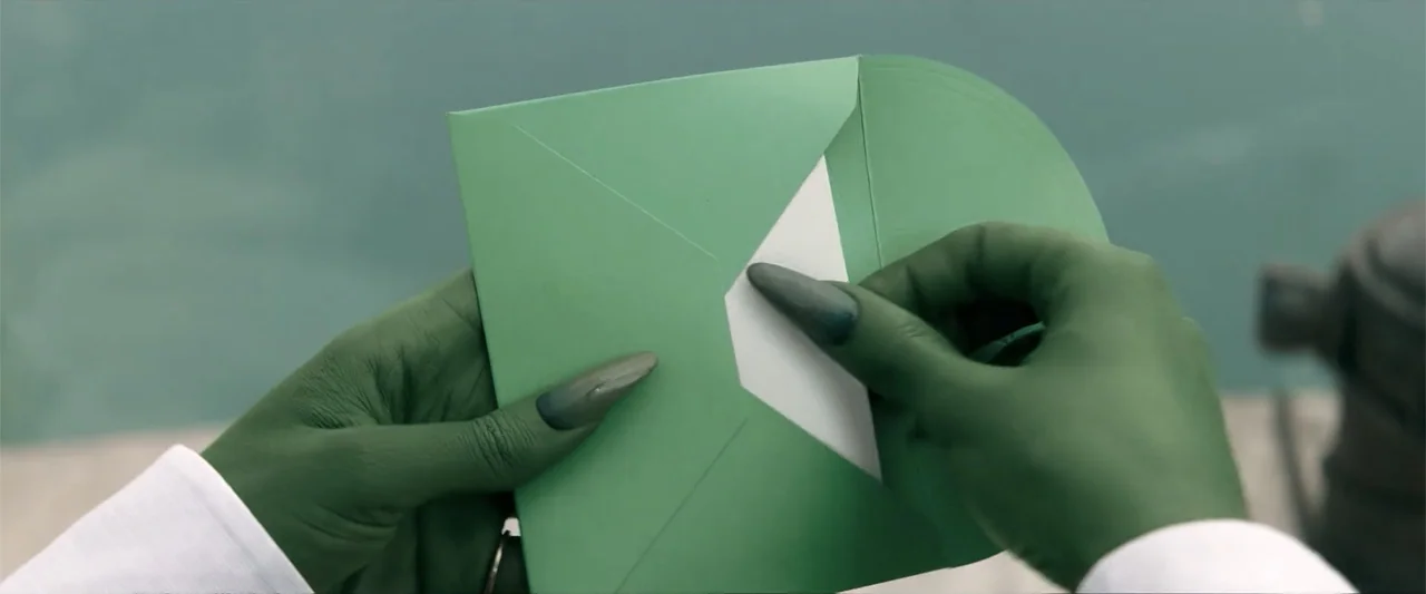 Wicked (1998) movie still: closeup — Hands painted green with long, dark, pointed fingernails hold an open green envelope. A white piece of paper…