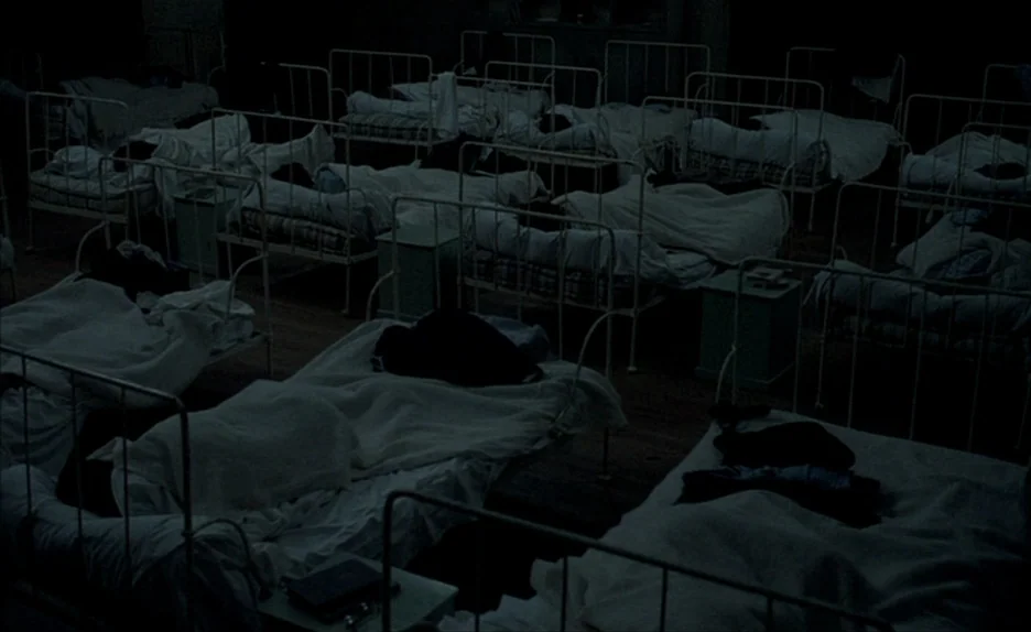 Au Revoir Les Enfants (1987) movie still: wide — A dark dormitory contains rows of metal bed frames covered with white linens. The room is filled…