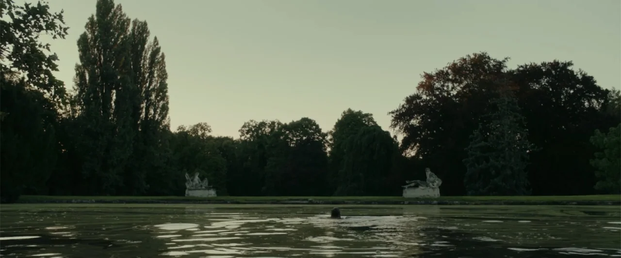 Peacock (2010) movie still: wide — An expanse of green murky water features stone statuary set upon a distant grassy bank. A single human head is…
