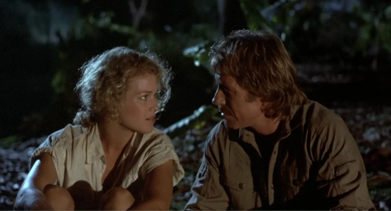 Firewalker (1986) movie still: medium — A nighttime outdoor setting with dense foliage and dark shadows in the background. A woman and man sit near…