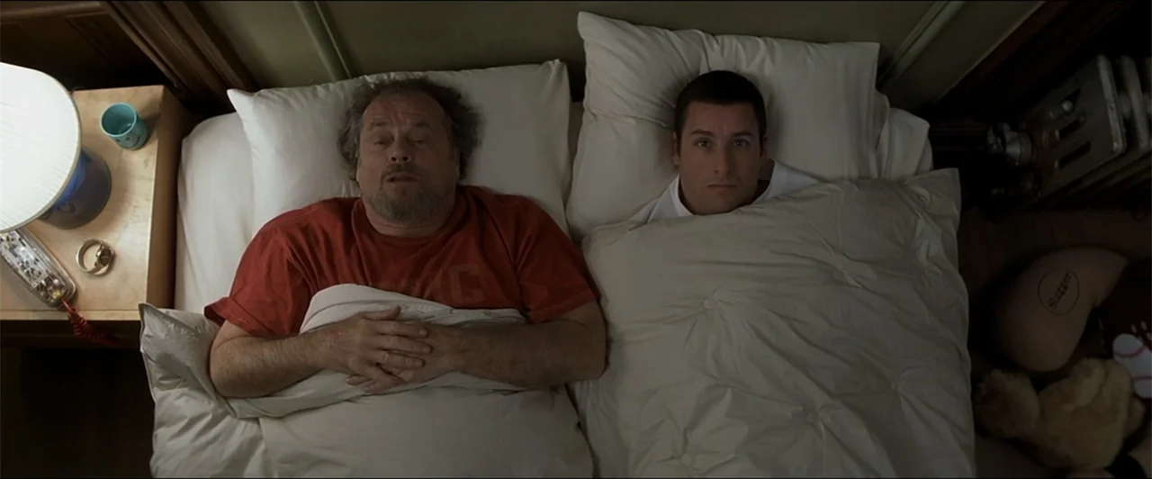 Anger Management (2003) movie still: medium — Two men lie together in a bed under white bedding in a dimly lit room. They both stare upwards at the…