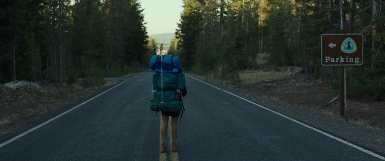 Wild (2014) movie still: fullbody — A quiet paved road runs through a dense pine forest during the daytime. A lone hiker carrying a large,…