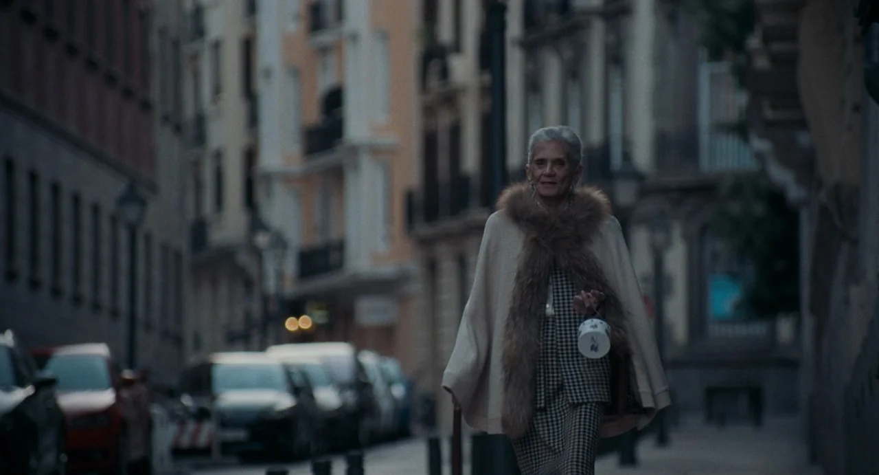 La Abuela (2021) movie still: fullbody — An elderly woman walks down a quiet city street lined with tall buildings. Parked cars are visible on the…