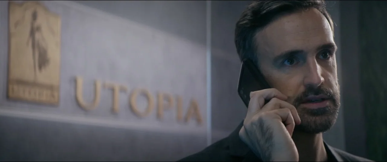 Utopia (2013) movie still: closeup — A sterile office interior features a grey wall with metallic gold lettering spelling Utopia. A man in a dark…