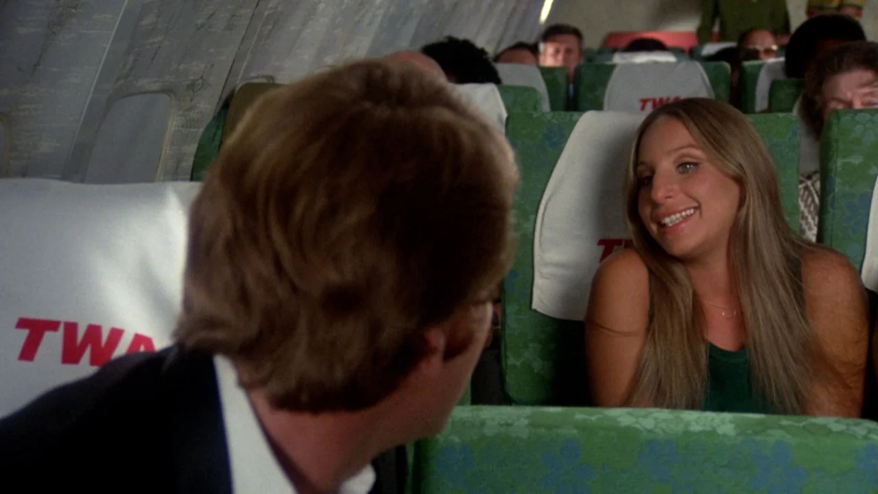 What’s Up, Doc? (1972) movie still: medium — An airplane cabin features rows of green patterned seats with TWA branded headrests. A woman with long…