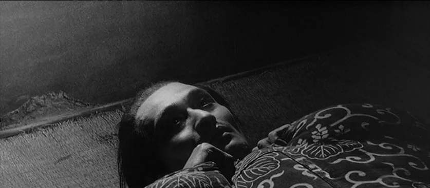 Red Beard (1965) movie still: closeup — A man lies on his side upon a woven tatami mat surface. The subject gazes upward into the darkness of the…