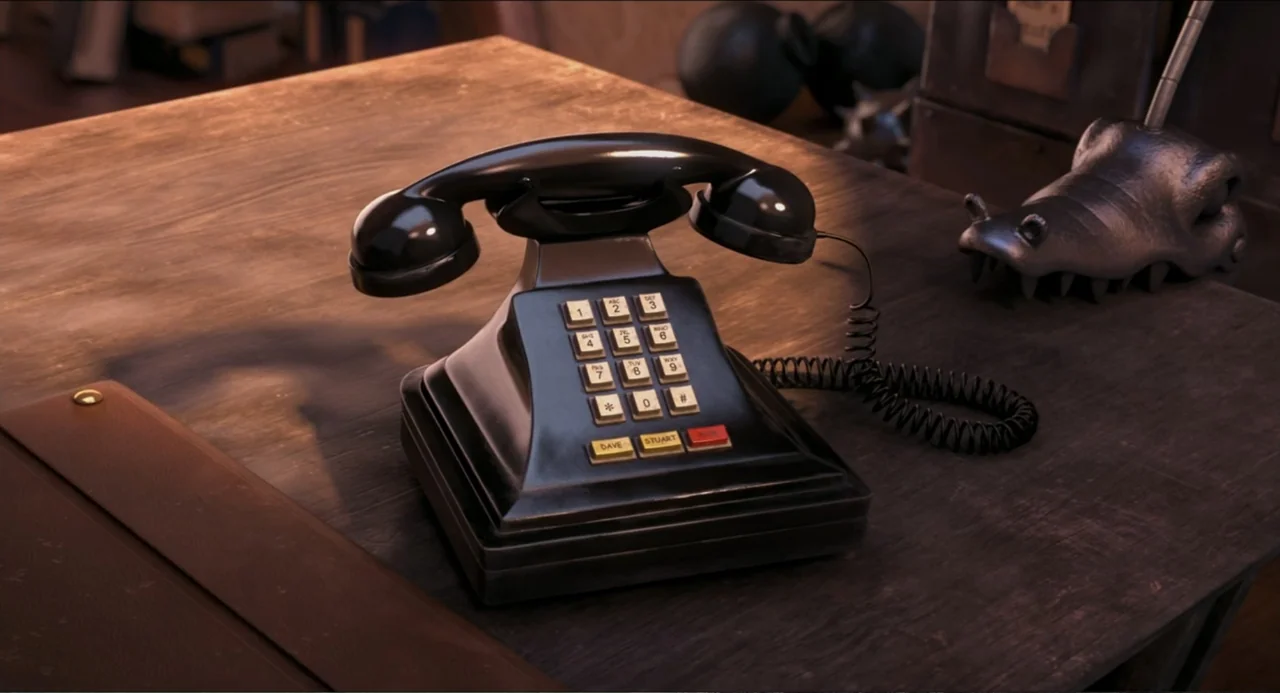 Despicable Me 2 (2013) movie still: closeup — A wooden desktop surface holds a black push-button telephone and a metal ornament. The telephone has…