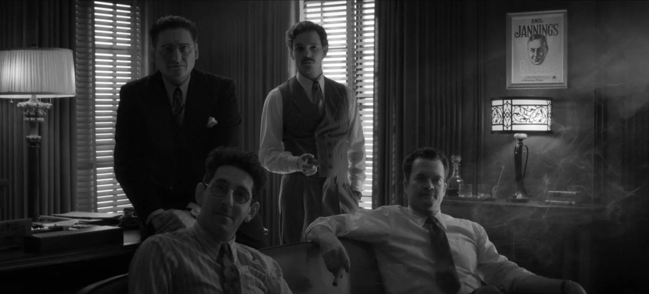 Mank (2020) movie still: medium — A dark office setting features wood paneling and window blinds filtering daylight. Four men in period attire…