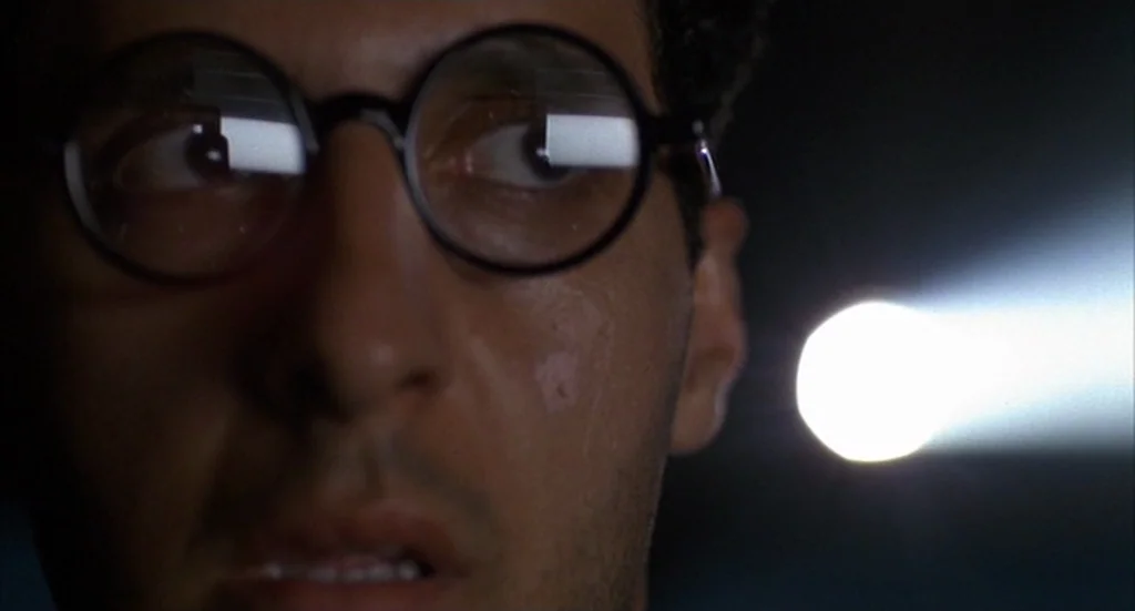 Barton Fink (1991) movie still: closeup — A dark, empty space illuminated by a distant, bright light source. A man in eyeglasses watches a screen in…