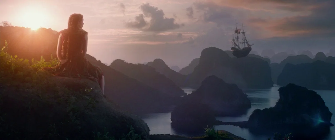 Pan (2015) movie still: wide — A woman sits on a grassy cliff overlooking a tropical bay with limestone islands. A sailing ship floats in the air…