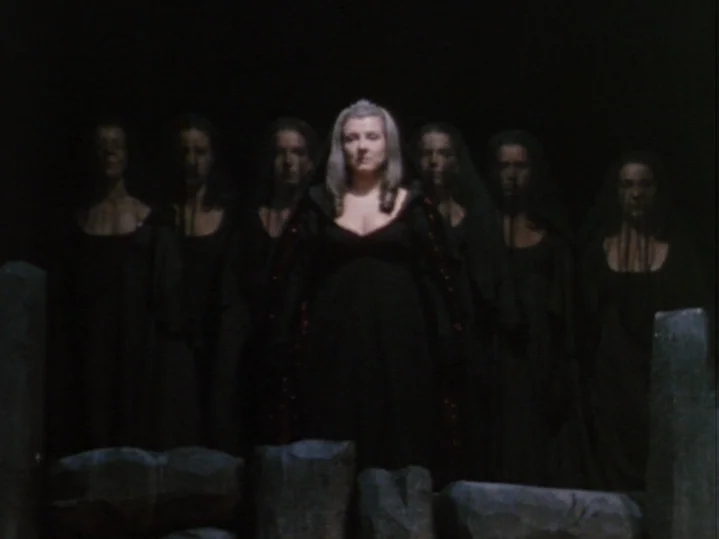 The Magic Flute (1975) movie still: medium — A group of women in black robes stands behind low stone pillars at night. The central woman with light…