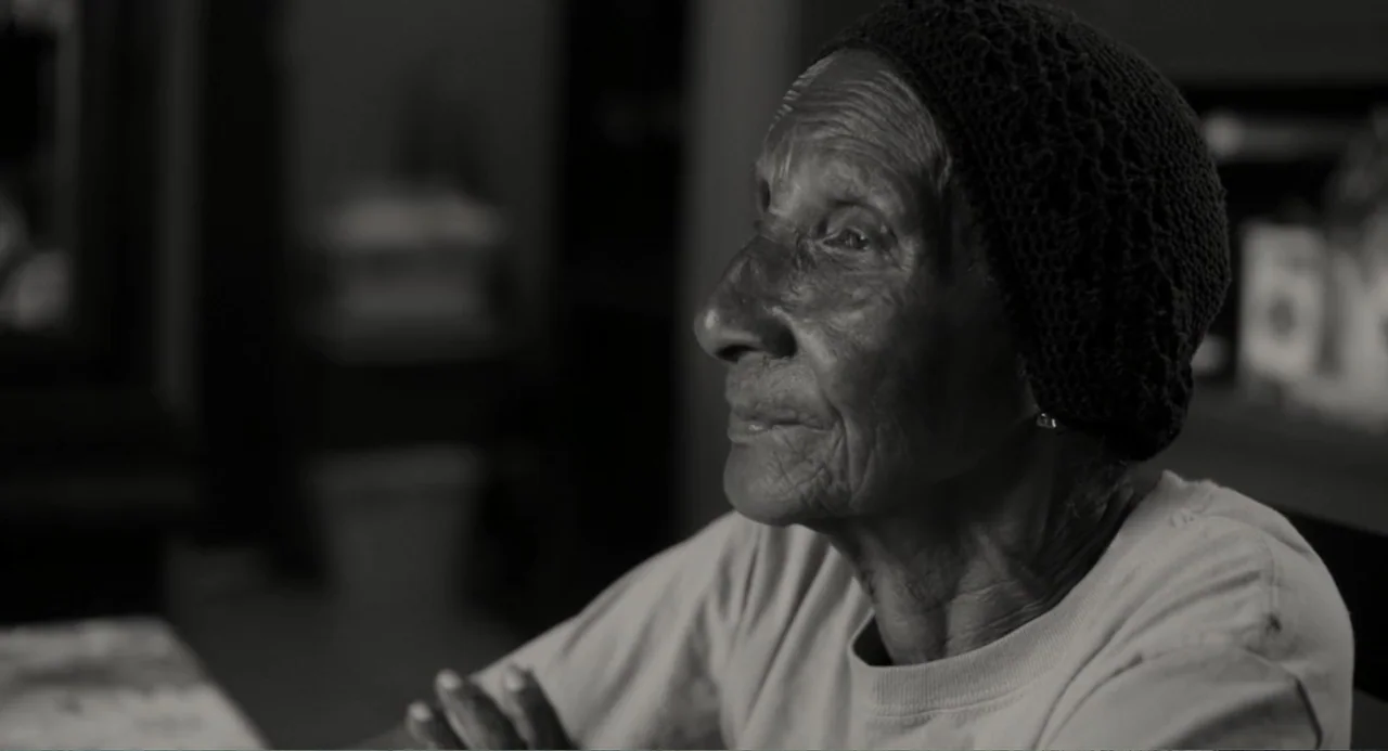 What You Gonna Do When the World's on Fire? (2019) movie still: medium — An interior space features an elderly woman seated against a dimly lit…