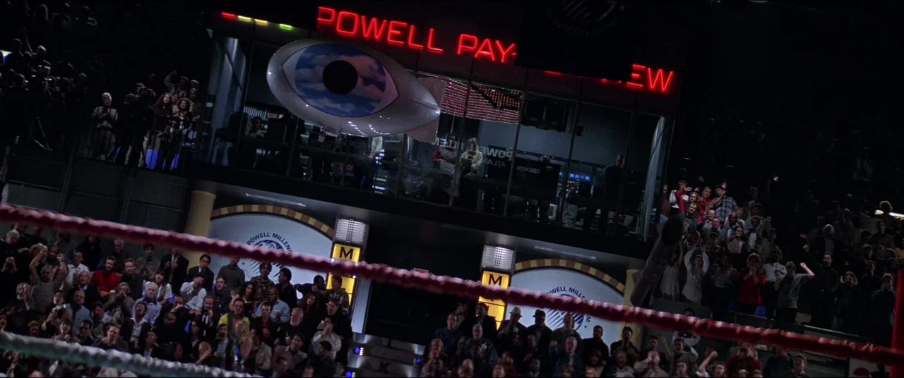 Snake Eyes (1998) movie still: wide — A multi-level indoor sports venue features a prominent blimp-shaped display with an eye logo. A large audience…
