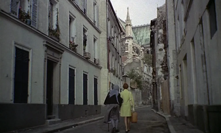 Nuits Rouges (1974) movie still: fullbody — A narrow European city street flanked by multi-story residential buildings with tall shutters. Two women…