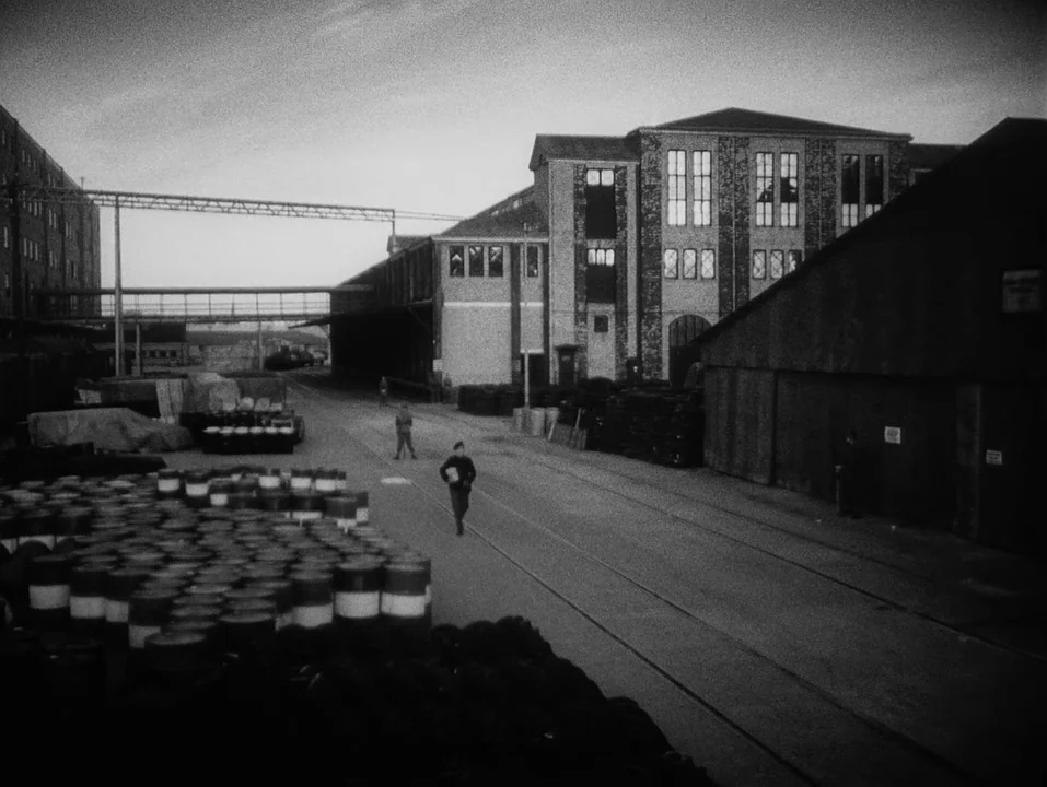 A Ship Bound for India (1947) movie still: wide — An industrial facility features large brick warehouses and an expansive paved outdoor yard. A lone…