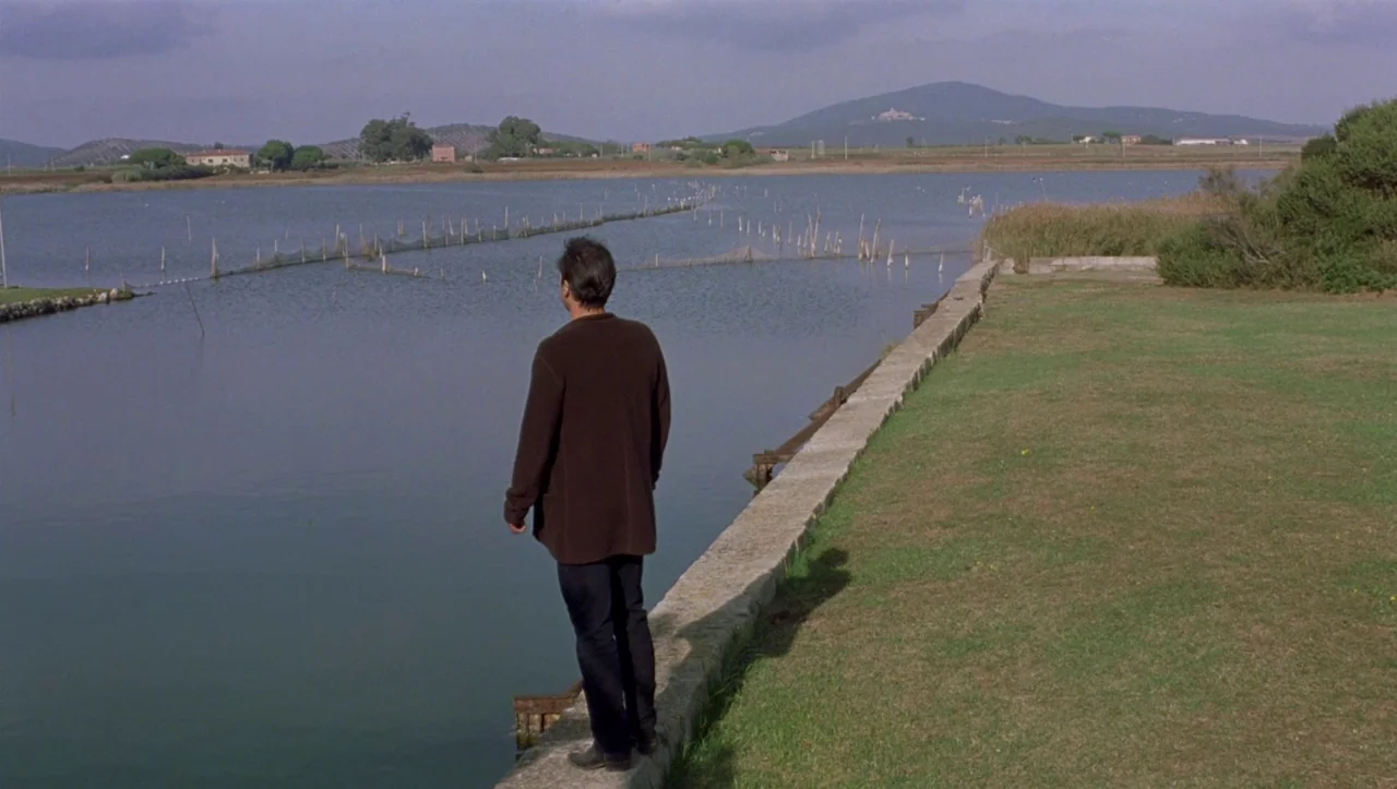 Eros (2004) movie still: wide — A man stands on a stone ledge beside a quiet lake with netting. Distant rolling hills are visible under a partly…