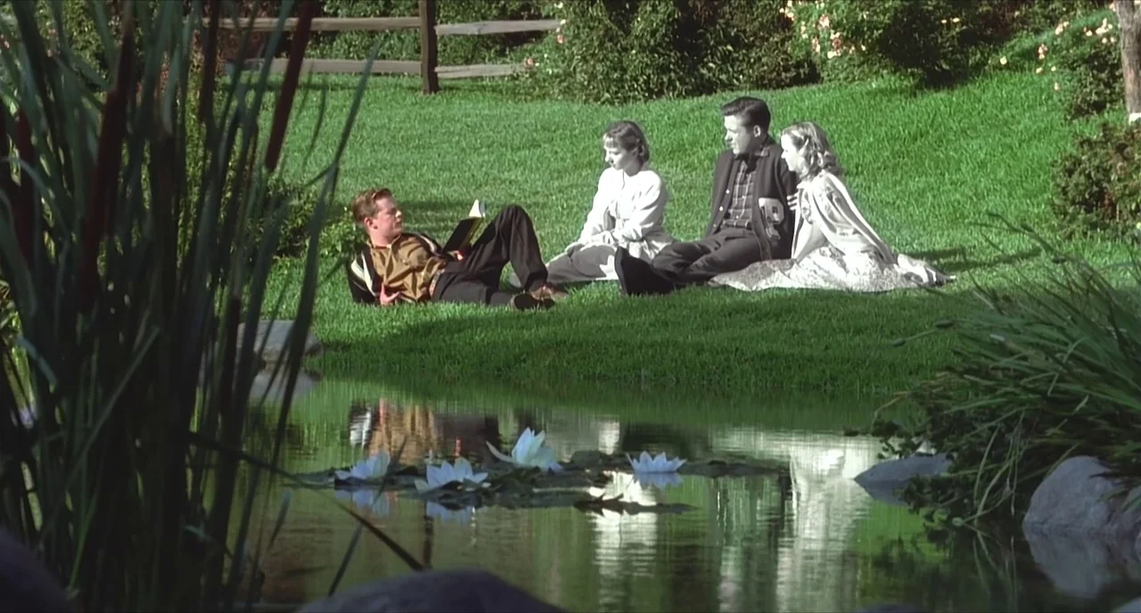 Pleasantville (1998) movie still: wide — Four young adults sit together on a manicured grassy hill beside a small pond. Water lilies bloom on the…