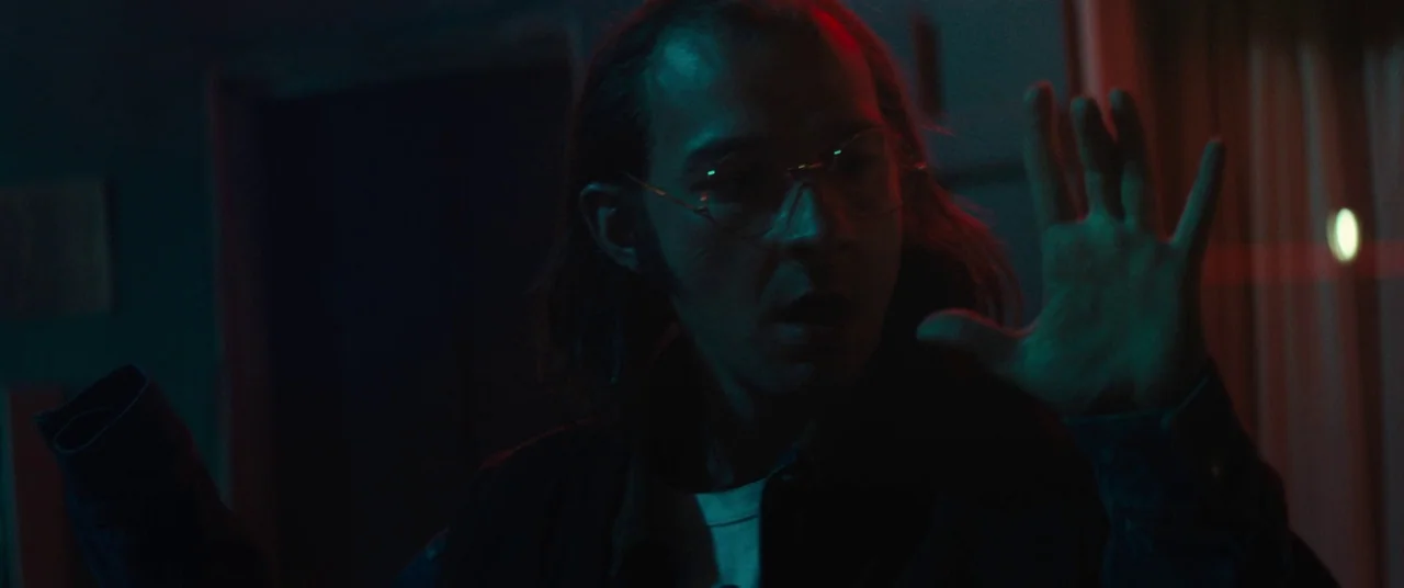 Honey Boy (2019) movie still: closeup — A dark indoor space is lit by high-contrast red and teal neon lighting. A man with long hair and glasses…