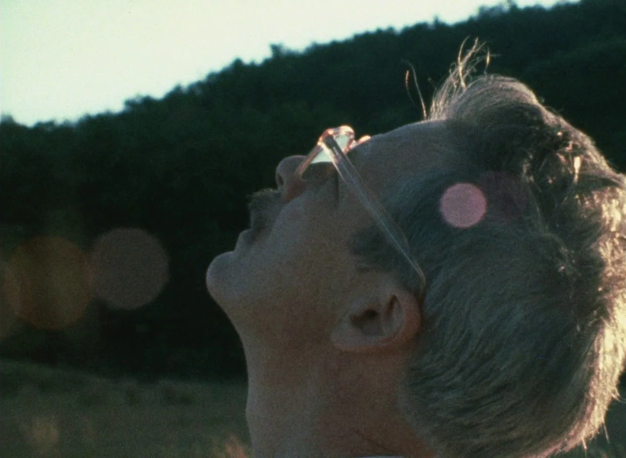 Wanda (1970) movie still: closeup — A field at golden hour with a dark treeline in the blurred background. A man tilts his head back while sunlight…