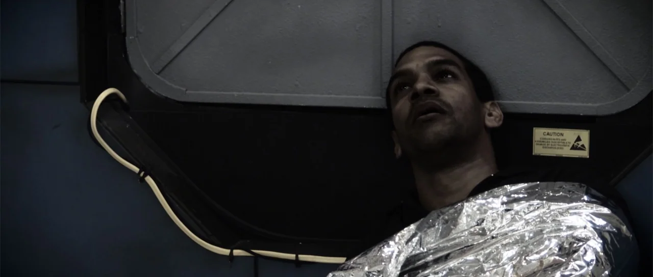 Astronaut: The Last Push (2012) movie still: closeup — A dark, cramped compartment features gray metal wall panels and industrial warning labels. A…