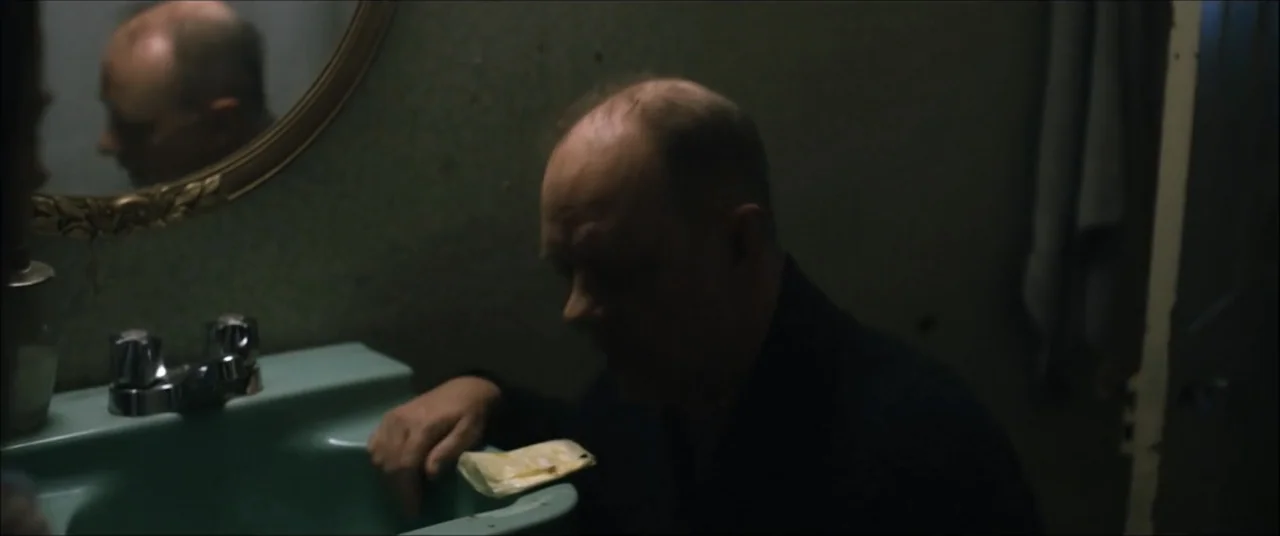 I'll Take Your Dead (2018) movie still: medium — A dimly lit bathroom features a vintage green basin and a wall-mounted mirror. An elderly man with…