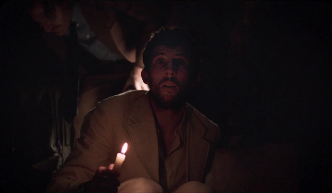 La Chimera (2023) movie still: medium — A dark, undifferentiated interior space with deep shadows obscuring the surroundings. A man with stubble…