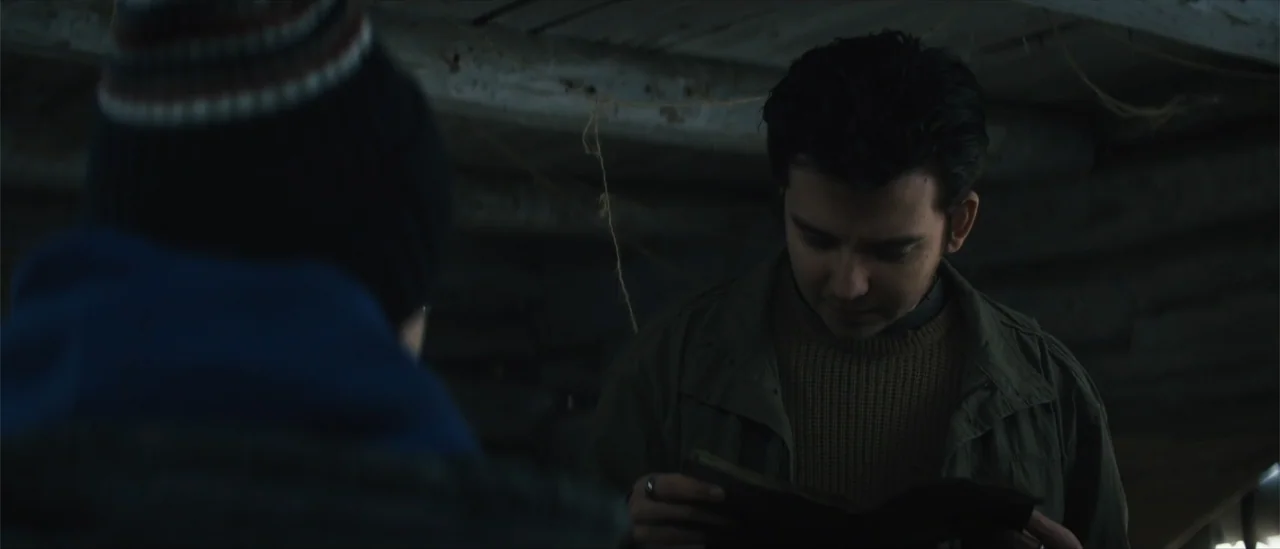 All Fun and Games (2023) movie still: medium — A dark wooden shed interior with rough hewn walls and dimly lit corners. A young man sits holding an…