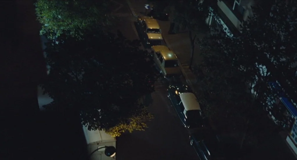 Something in the Air (2012) movie still: establishing — A nighttime view of a street with several cars parked along the curb. Large trees overhang…
