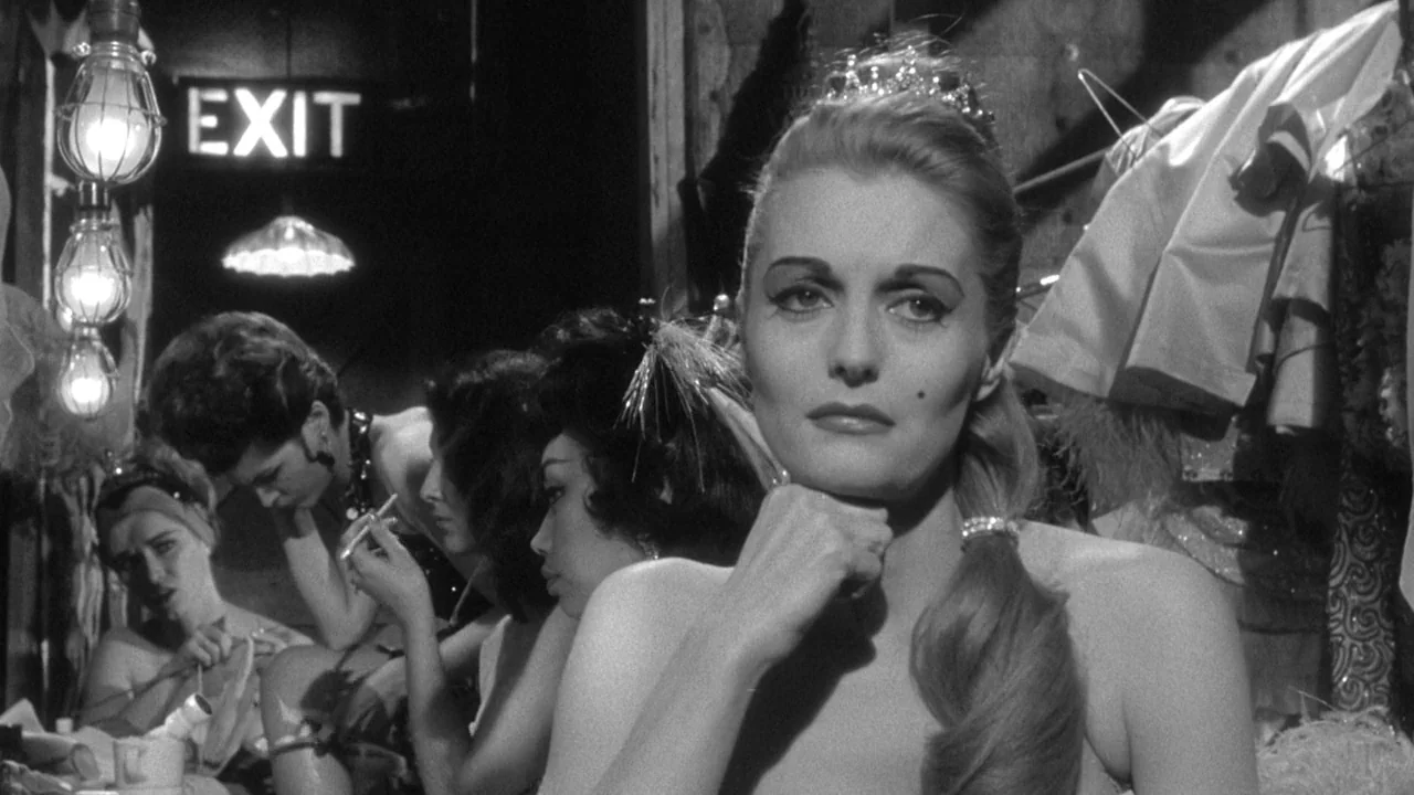 Shock Corridor (1963) movie still: medium — A cluttered backstage area with a lit exit sign and hanging costume racks. The primary woman sits in the…
