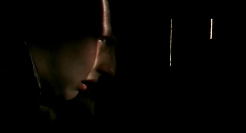 28 Days Later (2002) movie still: closeup — A dark, empty space frames a man in profile within a singular slice of light. The subject gazes ahead…