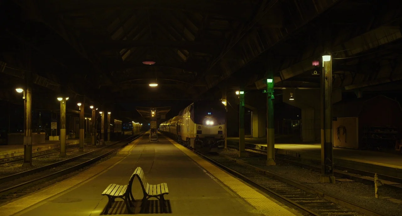 Last Flag Flying (2017) movie still: wide — A night view of a quiet rail platform with rows of overhead lights. An Amtrak train is stationary on the…
