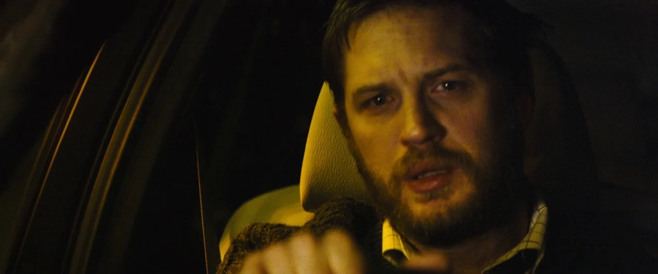 Locke (2013) movie still: closeup — The interior of a moving vehicle at night with dark surroundings. A man with a beard sits in the driver seat…