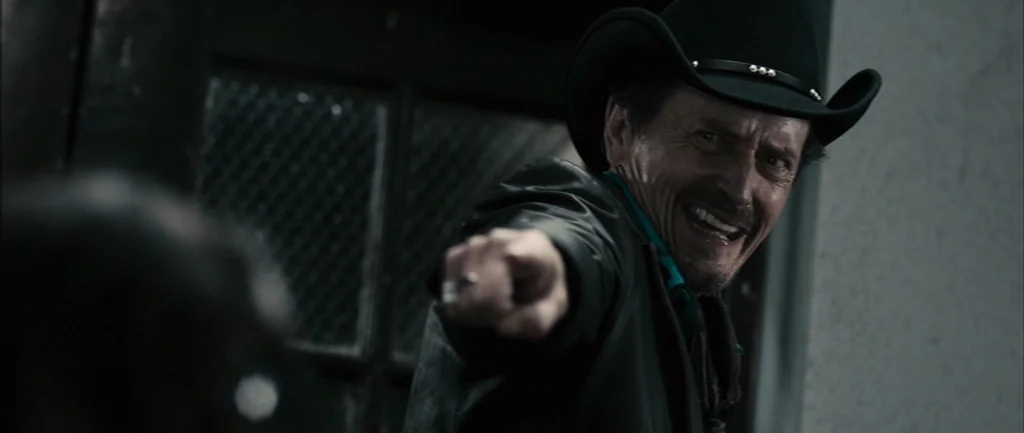Pontypool (2008) movie still: medium — A dark indoor space features a wall with a metal-grated window pane. A man in a cowboy hat points a finger…
