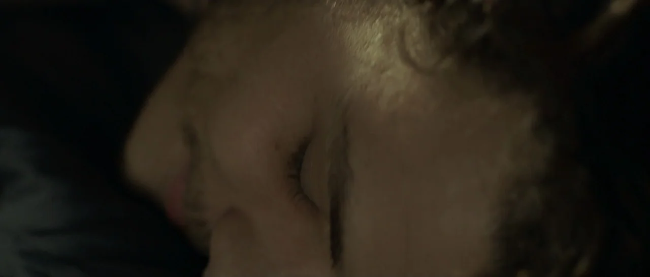 NY84 (2016) movie still: closeup — A dark, cramped interior space shows a person resting in deep shadow. The focus is tightly cropped on the side of…