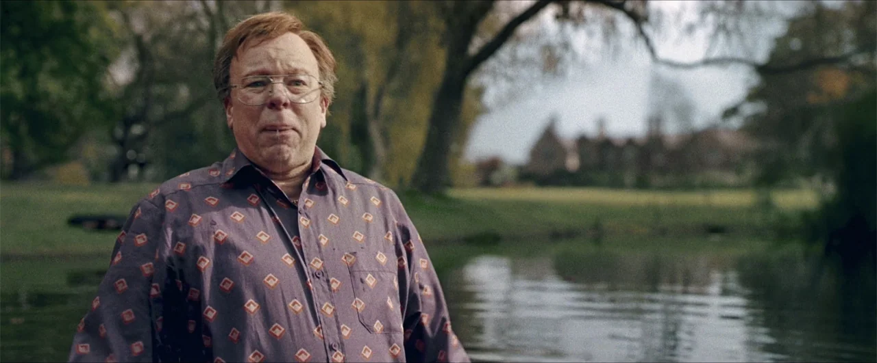 Better Man (2024) movie still: medium — A man stands in a murky lake with a country estate visible in background. The man in a patterned shirt…