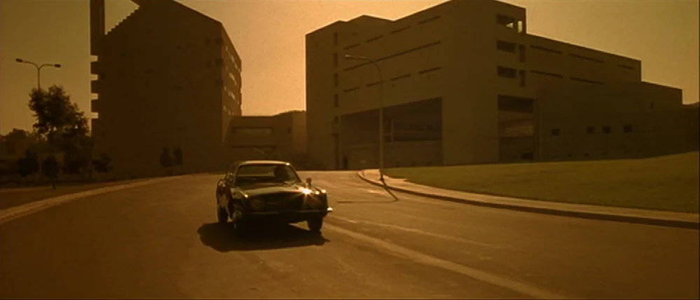 Gattaca (1997) movie still: wide — A dark sedan travels on a curved paved road between two large brutalist concrete buildings. The late afternoon…