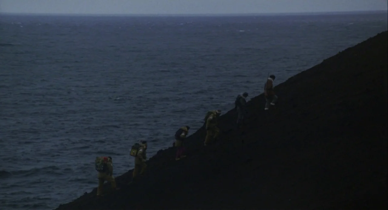 Godzilla vs. Mechagodzilla II (1993) movie still: wide — A group of five figures treks up a steep, black volcanic cliff side. The cold blue ocean…