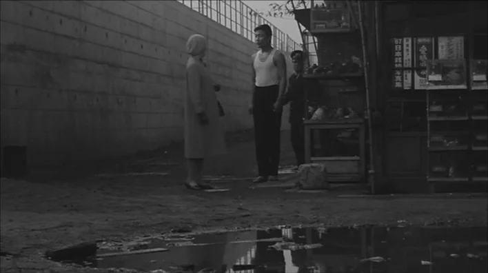 Death By Hanging (1968) movie still: fullbody — An urban area features a concrete wall alongside a small roadside vendor stand. A man in a tank top…