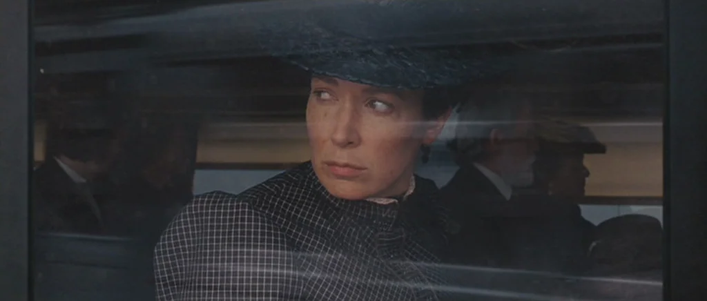 True Grit (2010) movie still: medium — A woman in a Victorian-era dress and hat gazes through a train window. Other blurred passengers are visible…
