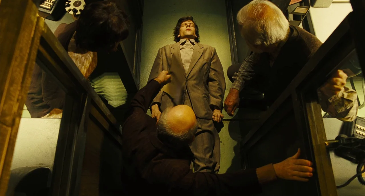 The Double (2013) movie still: fullbody — A narrow, dimly lit enclosure with dark walls features four people gathered together. A man in a suit lies…
