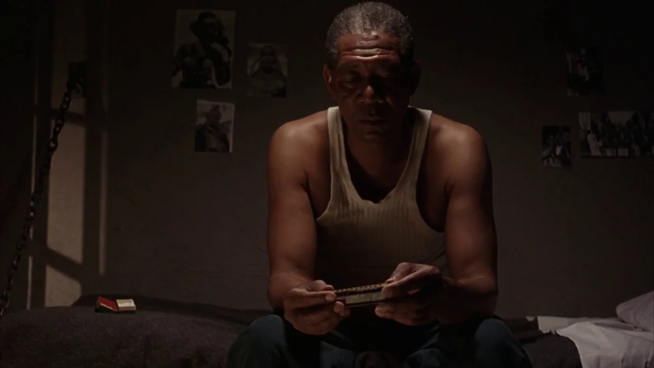 The Shawshank Redemption (1994) movie still: medium — A dim room has stone walls decorated with small, faded paper photographs. An elderly man sits…