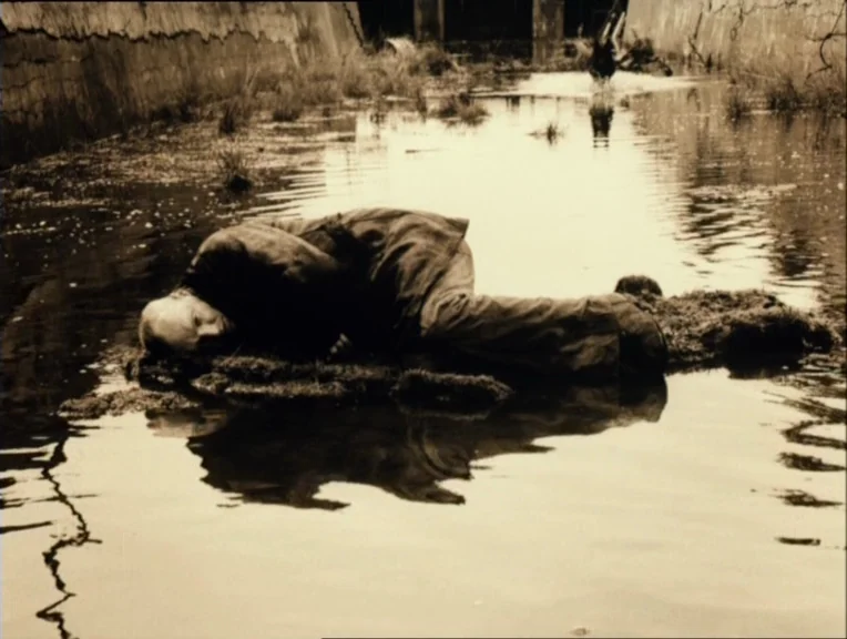 Stalker (1979) movie still: fullbody — A man lies on a small muddy island in a narrow, shallow water channel. The concrete banks of the channel rise…