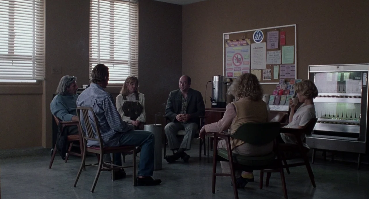 Touch (1997) movie still: wide — A windowless community center room with beige walls and a pinboard displaying posters. Six people sit on chairs…